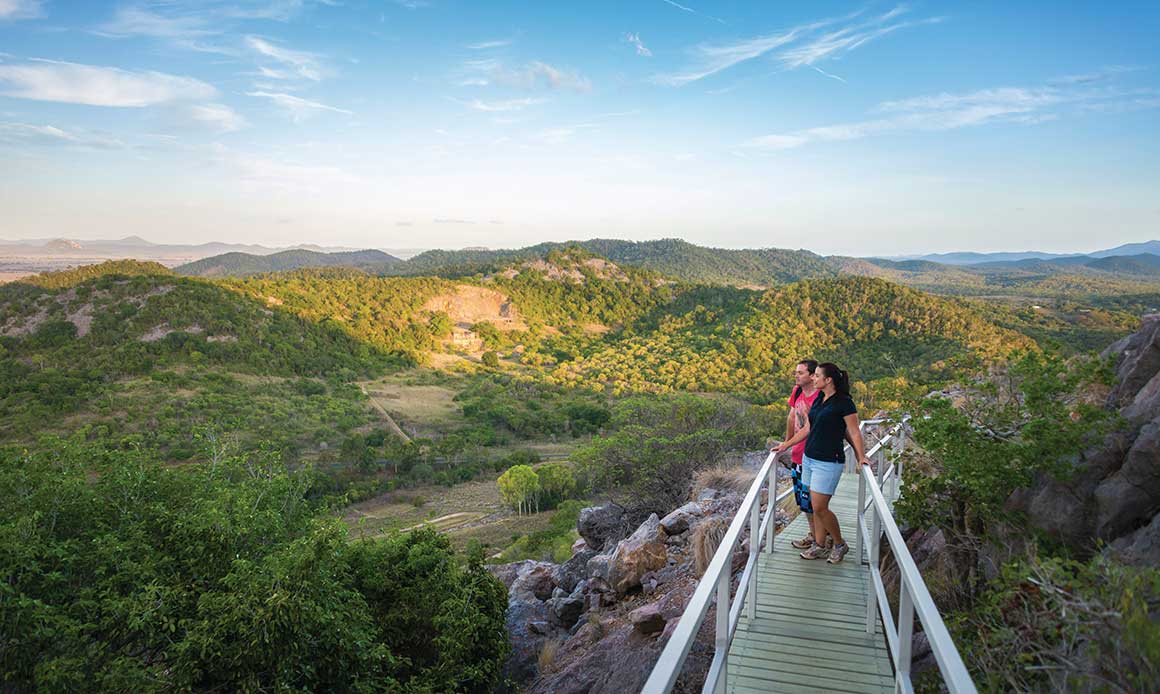 Couple stand on elevated walkway winding around a limestone outcrop, overlooking rolling limestone hills clad in dry rainforest. 