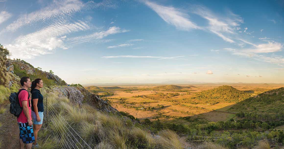 A couple stands at a lookout gazing over expansive landscape of flat plains and lumpy hills under big blue skies.