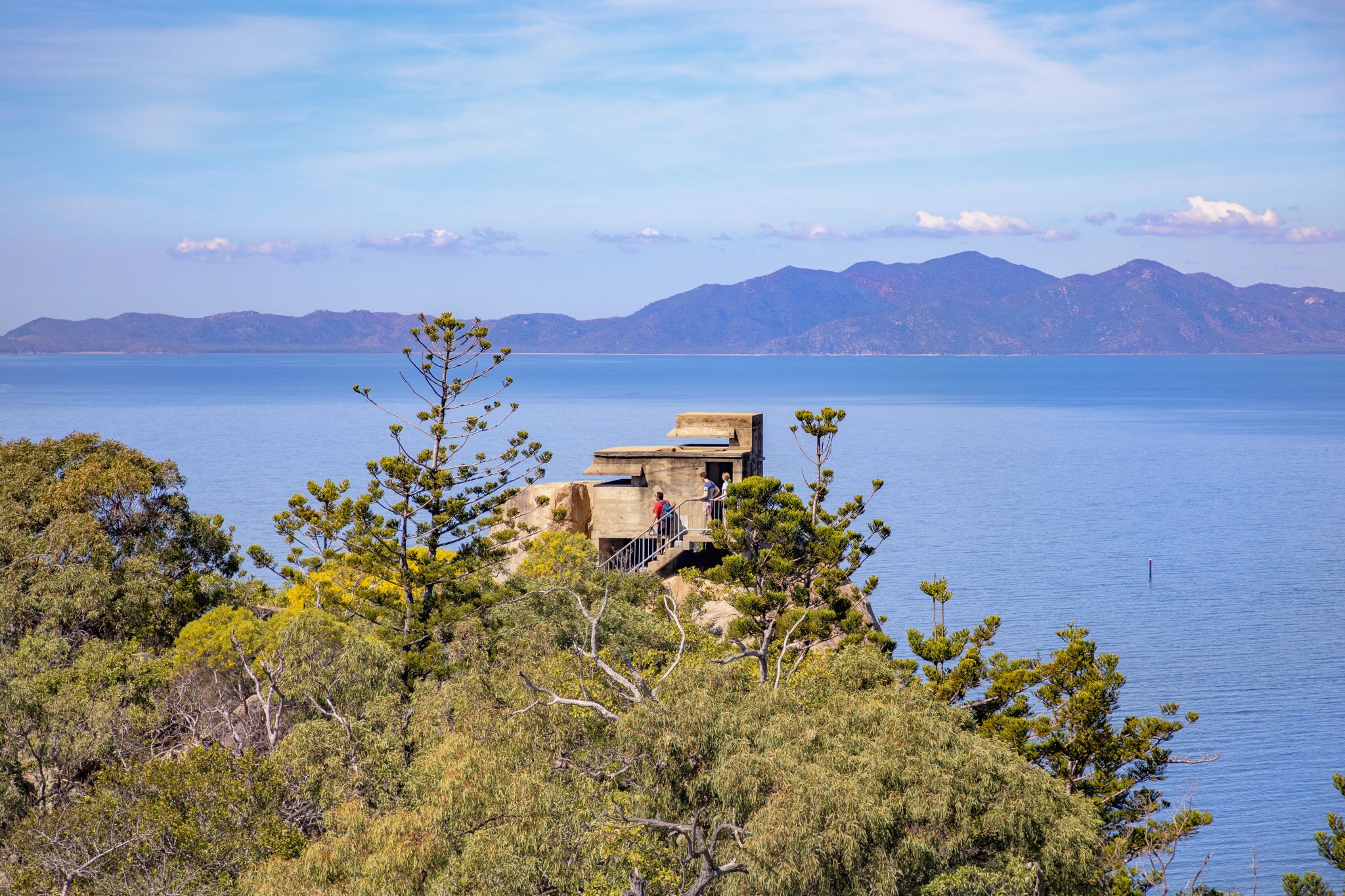 Three people climbing down stairs from an old concrete fort. Ocean and islands in the background and trees in the foreground.