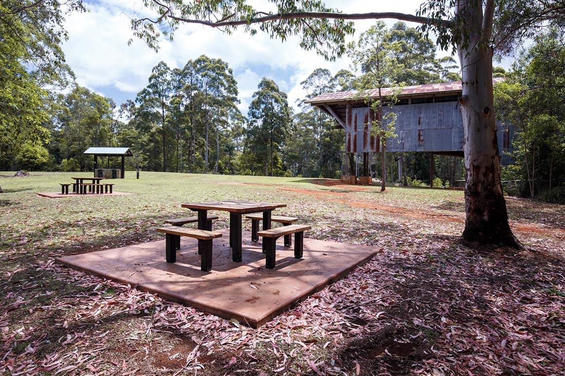 Picnic tables and picnic shelters are set in an open grassy space with an historic corrugated iron building and surrounding forest in the background. 