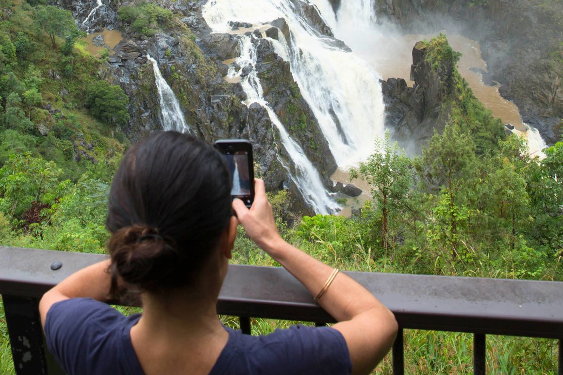 Person standing at lookout with arms on railing taking picture with phone of a waterfall.